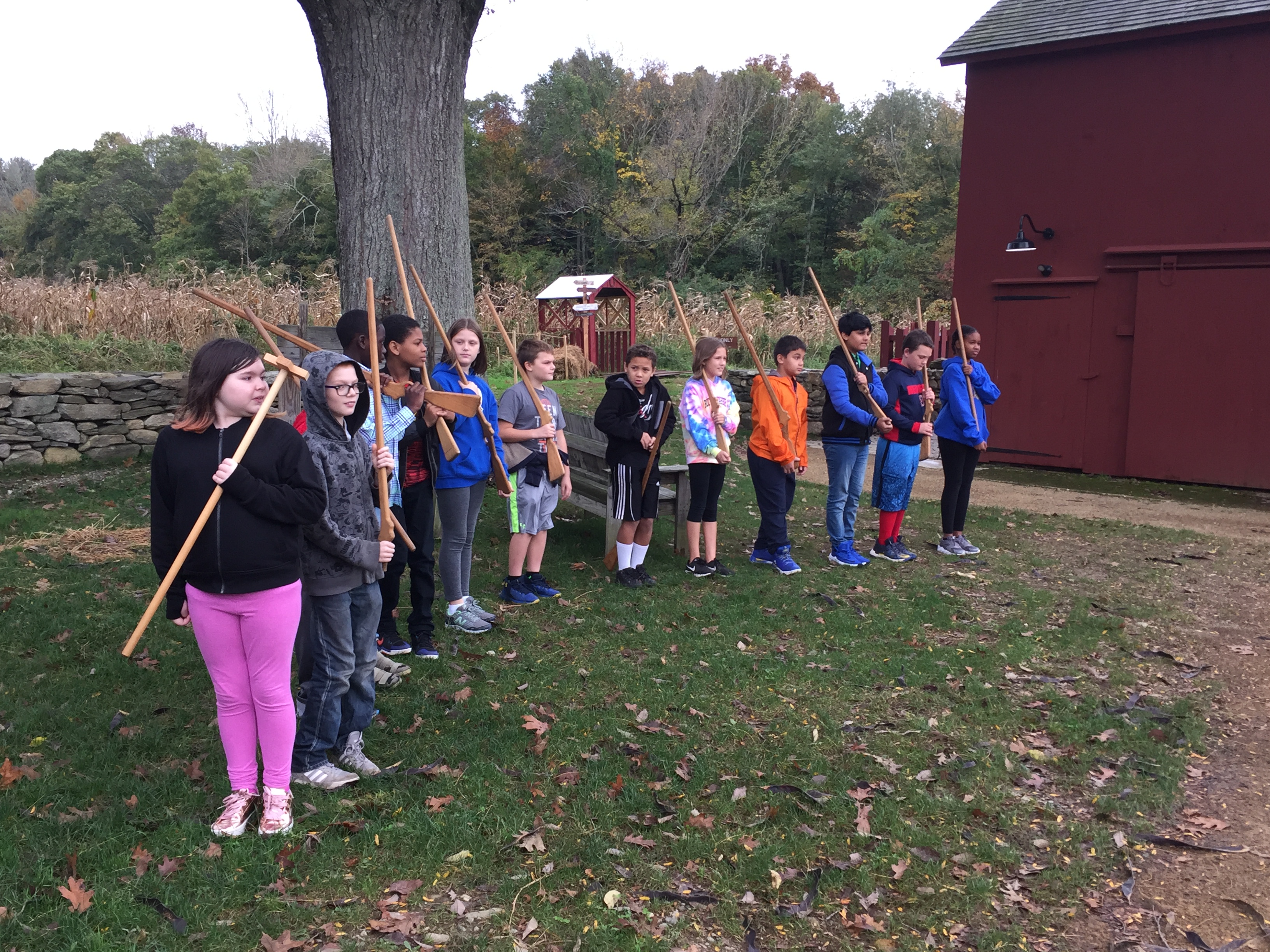 A group of children stands in a line outdoors, each holding a stick, with a red barn and trees in the background.