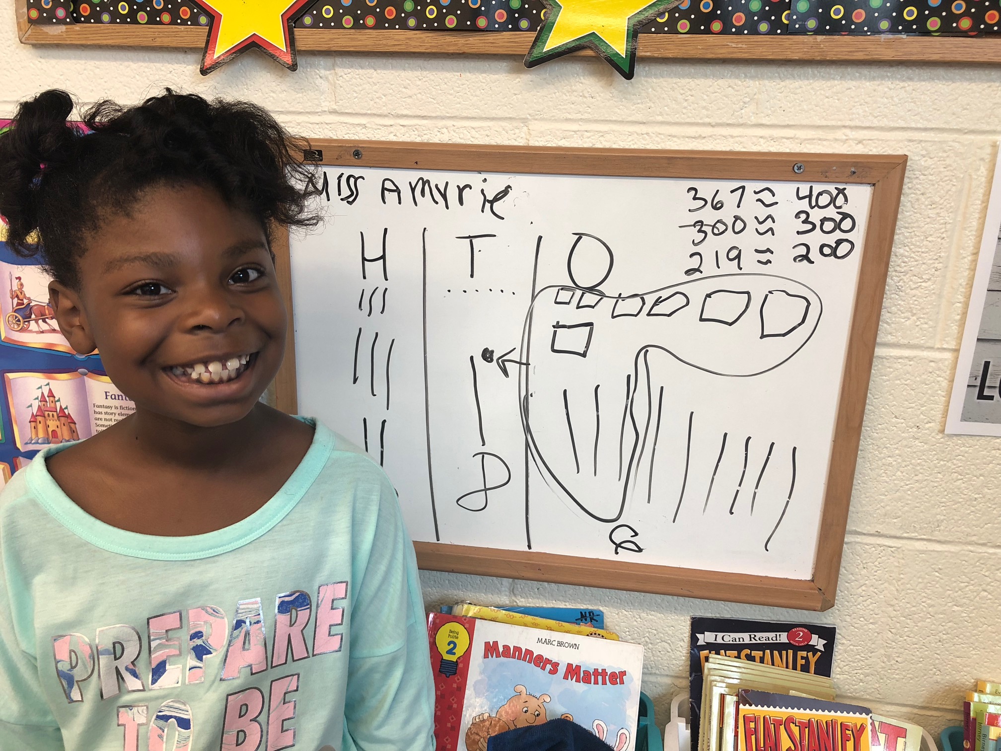 A smiling child stands next to a whiteboard displaying a drawing and some numbers, likely related to a classroom activity.