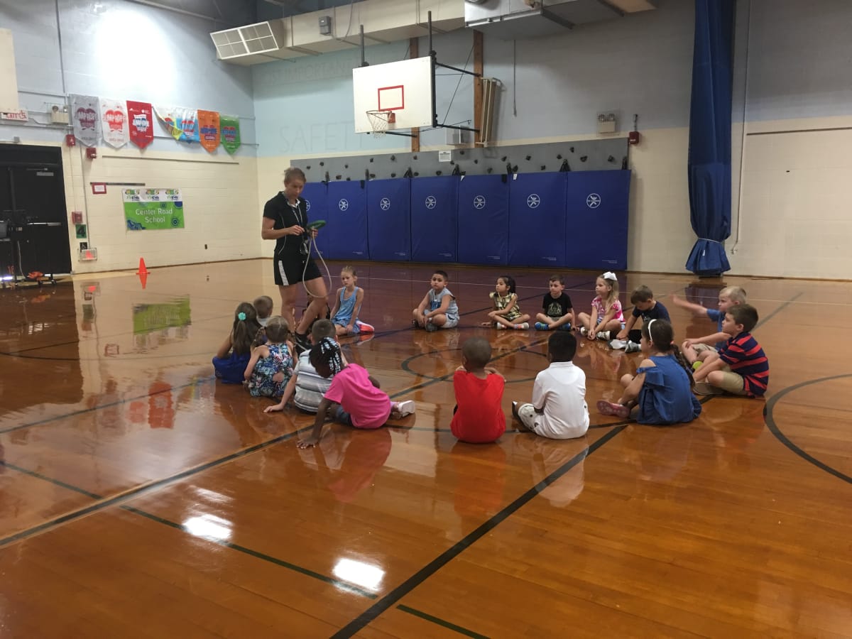 A group of children sitting in a circle on a gymnasium floor, listening to an adult instructor who is standing and speaking to them.