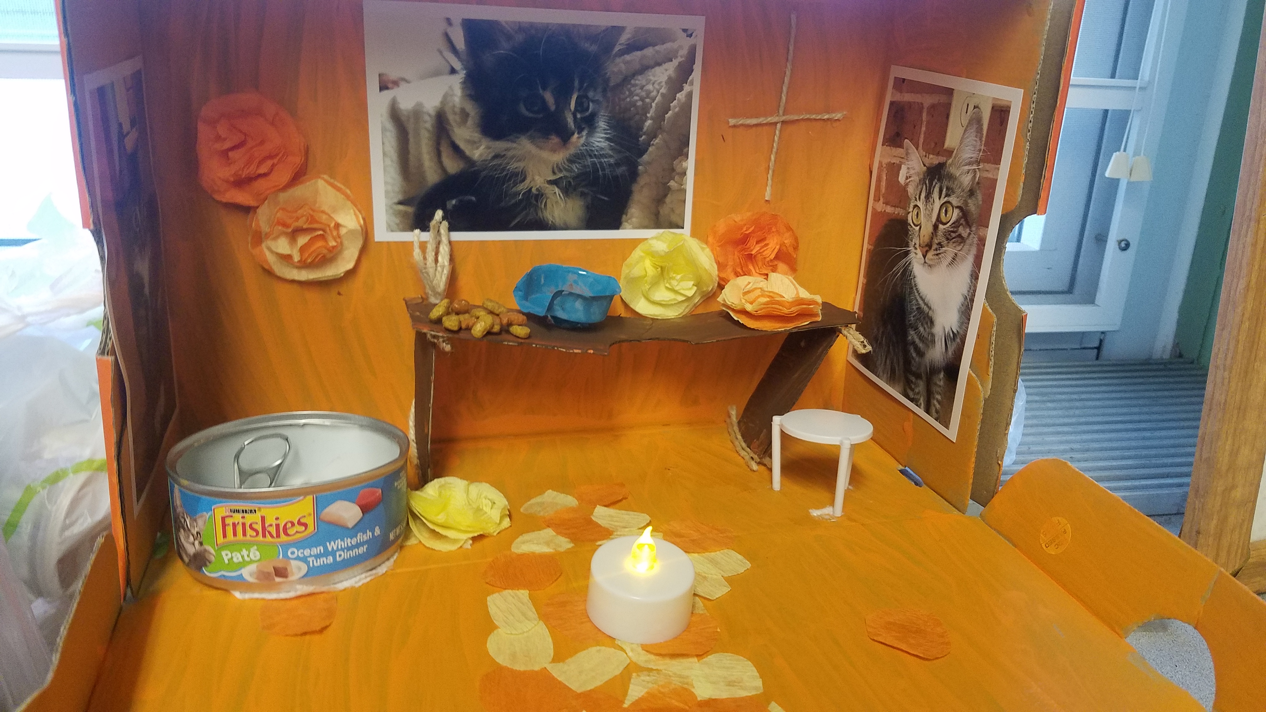 A small diorama featuring a table with a candle, a can of cat food, and decorative items, surrounded by photographs of cats on the walls.