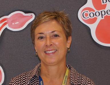 A smiling woman with short hair stands in front of a gray wall decorated with colorful shapes and text.
