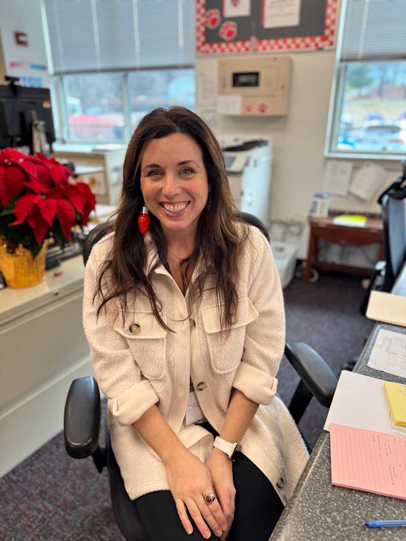 A smiling woman with long brown hair is sitting in an office chair, wearing a light-colored jacket, with a poinsettia plant in the background and various papers on the desk.