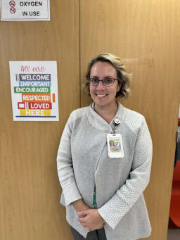 A smiling woman stands next to a sign that reads 'All are welcome, important, encouraged, respected, loved here,' with a notice above indicating 'Oxygen in use.'