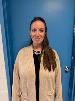 A woman with long brown hair is smiling while wearing a beige cardigan over a black top, standing in front of a blue wall and door.