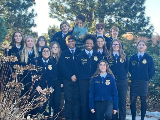 A group of students wearing blue jackets stands together outdoors, surrounded by trees and plants, smiling for the camera.