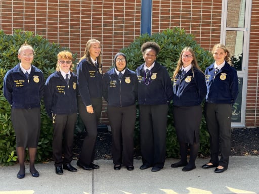 A group of seven individuals wearing navy blue jackets with emblems stands in front of a brick wall and green shrubs, smiling at the camera.