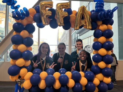 Four students pose together in front of a balloon arch decorated with blue and yellow balloons, with the letters 'FFA' prominently displayed above them.