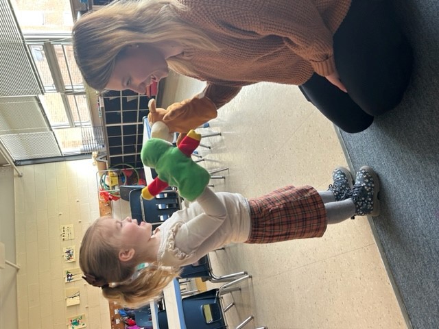 A young girl wearing a white top and plaid skirt playfully interacts with a woman, using a colorful puppet to mimic feeding her a drink in a classroom setting.