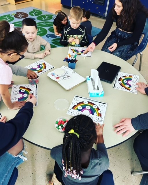 A group of children and adults are gathered around a table, engaged in a coloring activity with rainbow-themed worksheets and art supplies.
