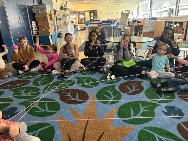A group of children and adults sit in a circle on a colorful carpet, holding strings that create a web-like pattern among them, suggesting a collaborative activity.