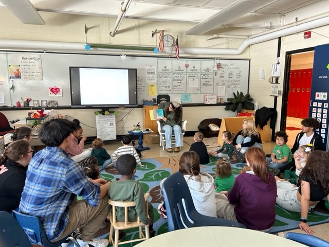 A group of children and adults are seated on a colorful rug in a classroom, while a person reads a book to them from a chair, with a whiteboard and a screen in the background.