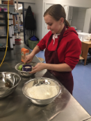A person in a red hoodie and apron is mixing ingredients in a bowl while standing at a kitchen counter with metal bowls and a workspace in the background.