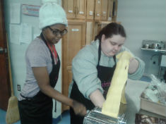 Two individuals are working together in a kitchen, with one person operating a pasta machine while the other assists by feeding dough into it.