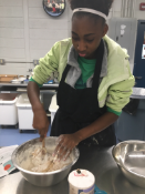 A person wearing an apron and a green jacket is stirring a bowl of batter with a wooden spoon in a kitchen setting.