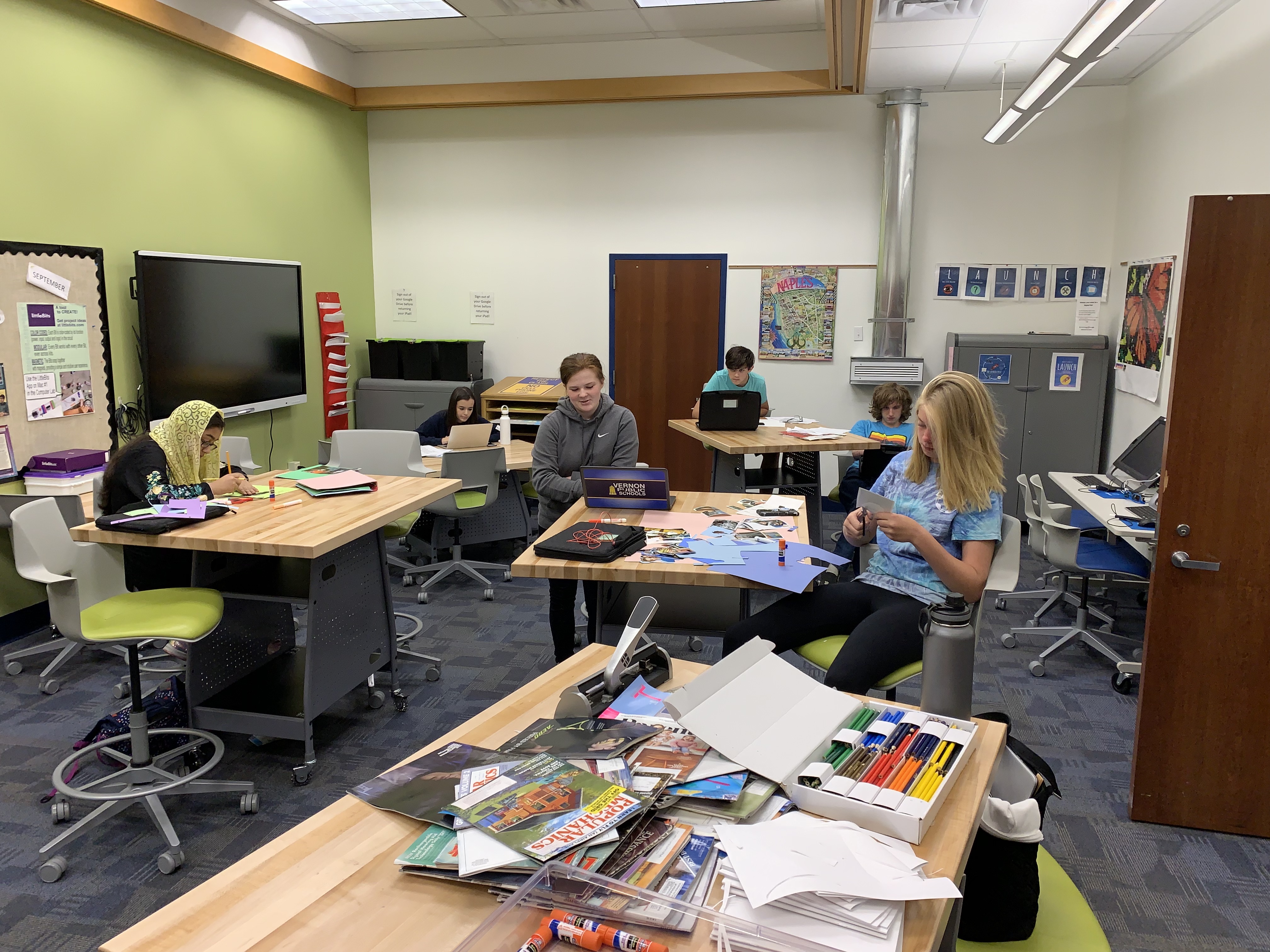 A classroom setting featuring several students engaged in various activities at tables filled with art supplies and materials, with green walls and modern furniture.