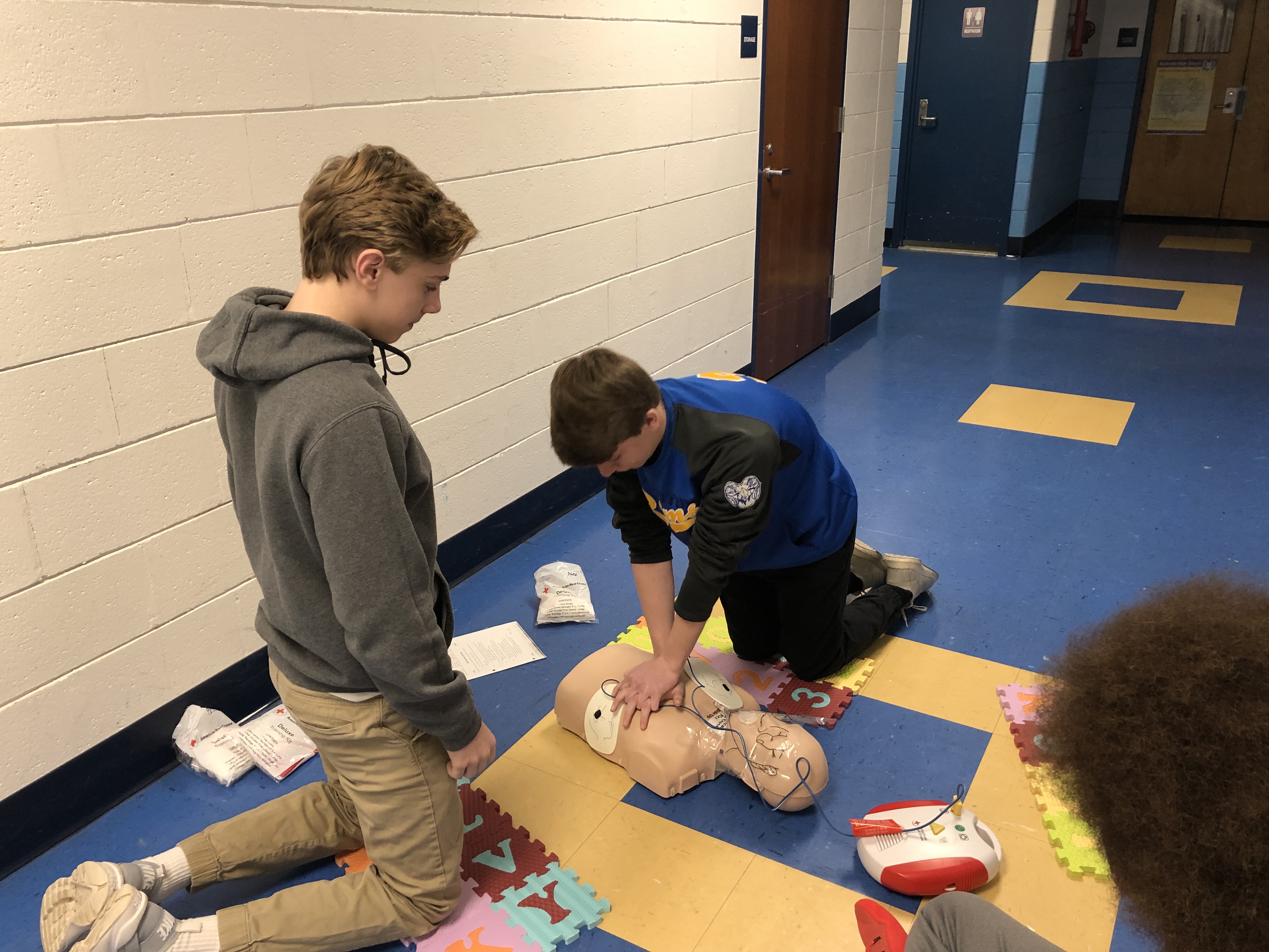Two students practice CPR on a training mannequin in a school hallway, with one student performing chest compressions while the other observes.