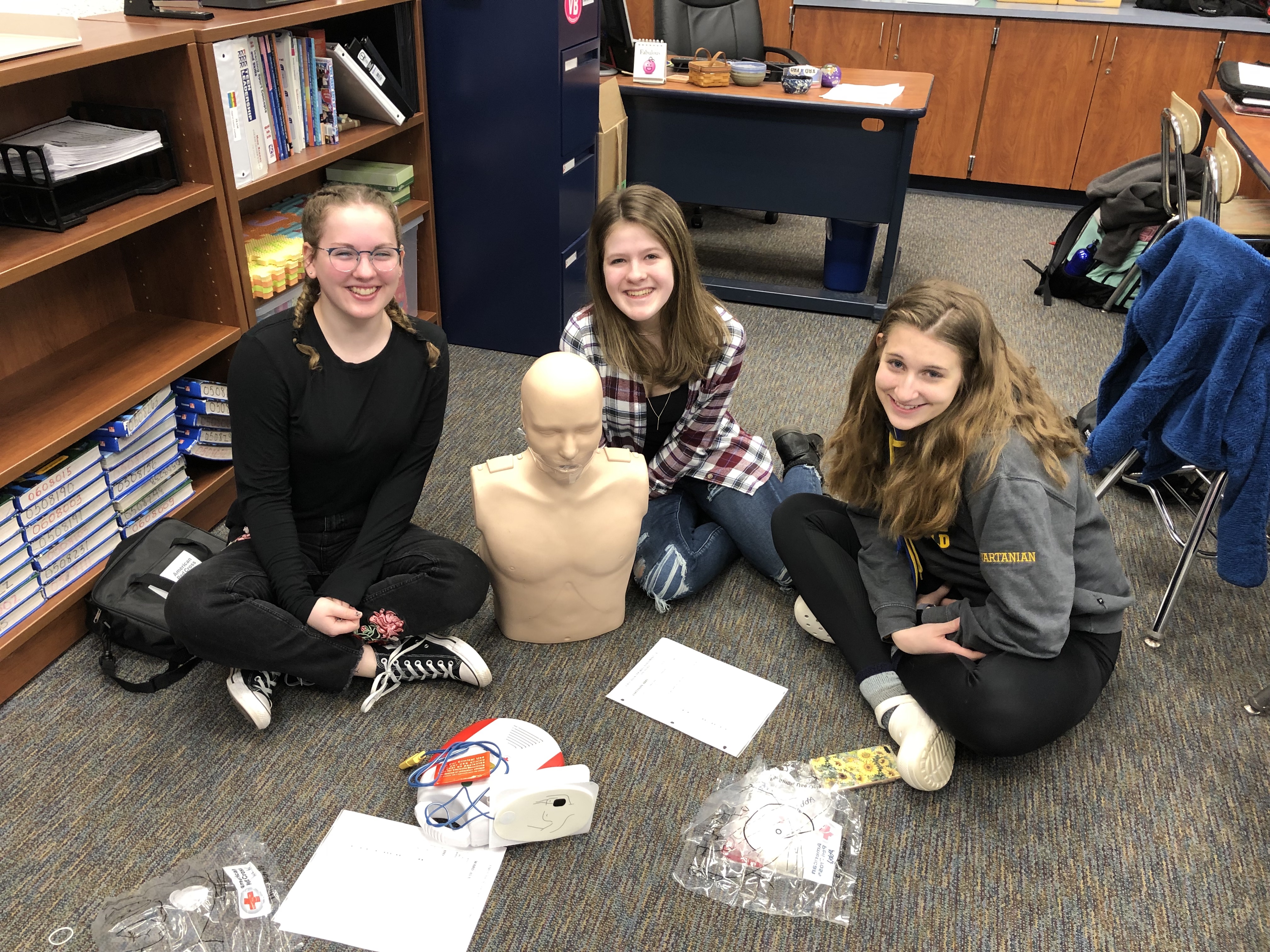 Three students sit on the floor in a classroom, smiling at the camera while surrounded by a mannequin and various educational materials.