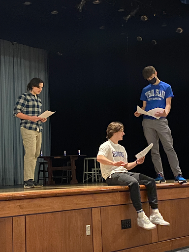 Three students are rehearsing a scene on stage, with one sitting on the edge of the stage and two standing, all holding scripts.