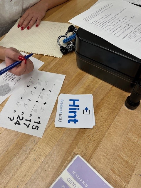 A person's hand holding a pen is seen above a game sheet with numbers and a card labeled 'Hint' on a wooden table, alongside a black box and some papers.