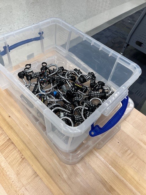A clear plastic container filled with numerous black and silver metal clips, placed on a wooden surface.