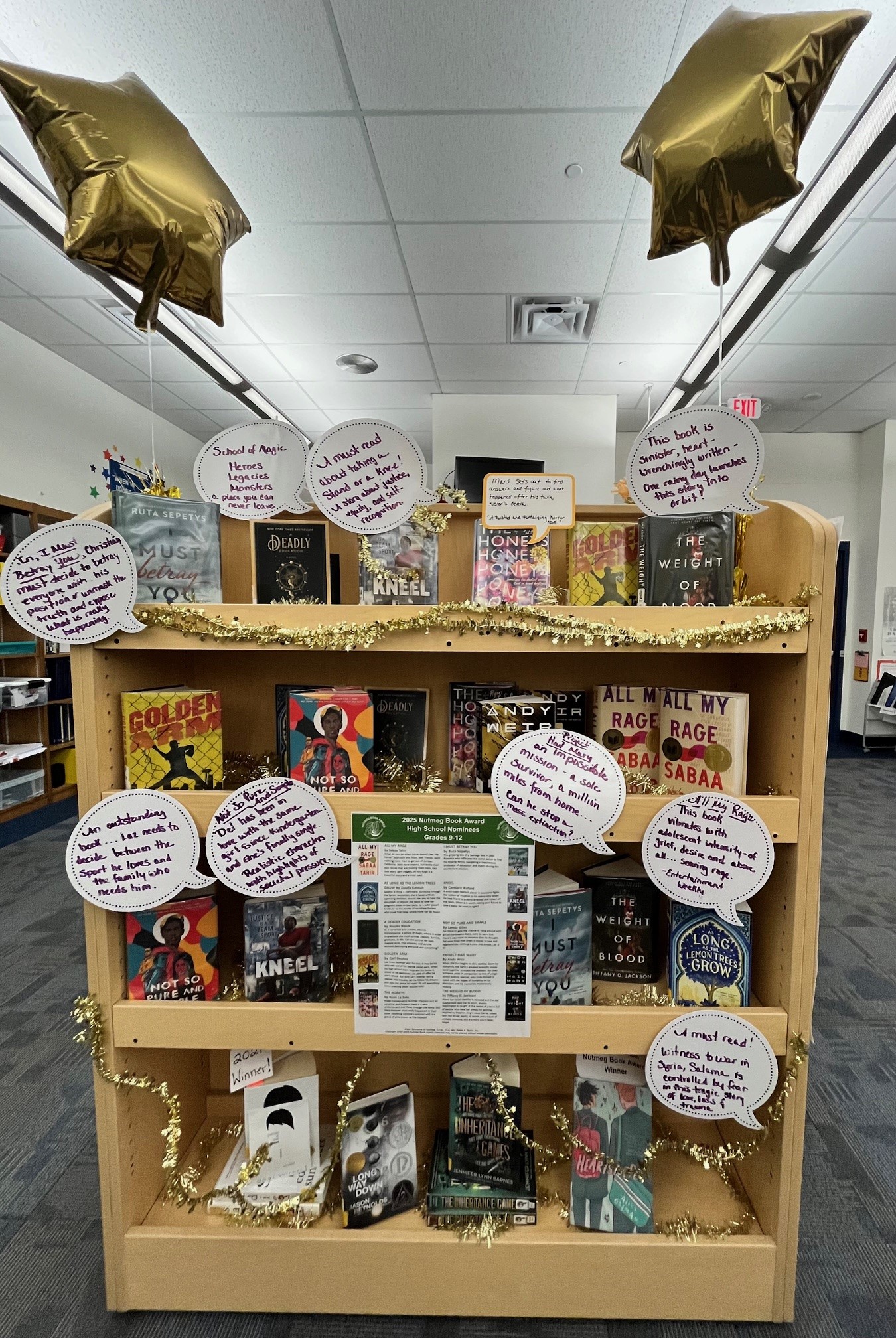 A wooden bookshelf displaying various books, decorated with gold balloons and circular signs featuring quotes or descriptions related to the books.