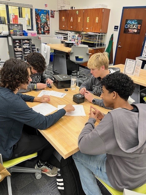 Four students are seated around a table in a classroom, engaged in a collaborative activity with papers and a small device in front of them.