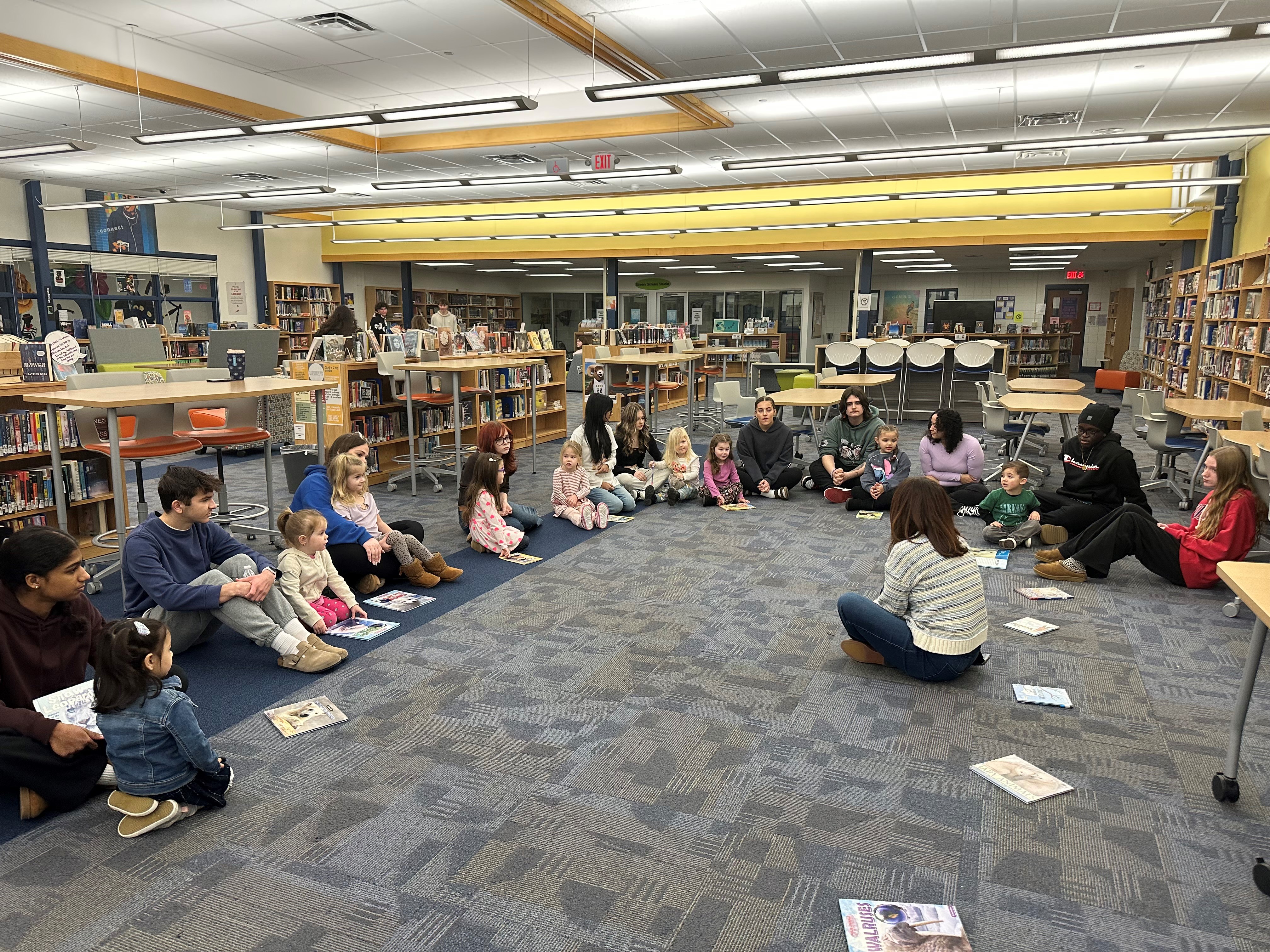 A group of children and adults are seated in a circle on the floor of a library, with books scattered around, as they listen to a person speaking in the center.