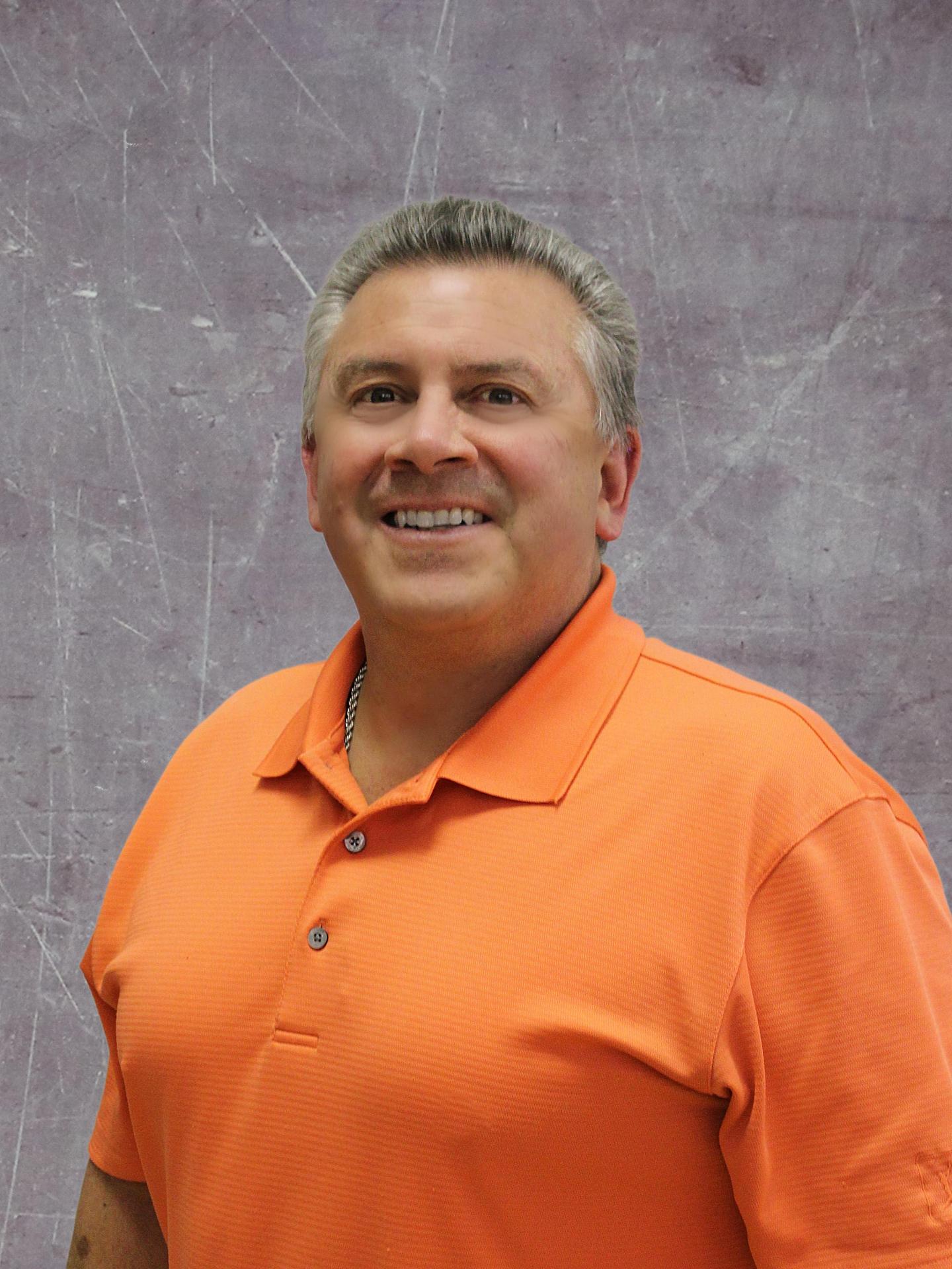 A smiling man with short gray hair wearing an orange polo shirt stands against a textured gray background.