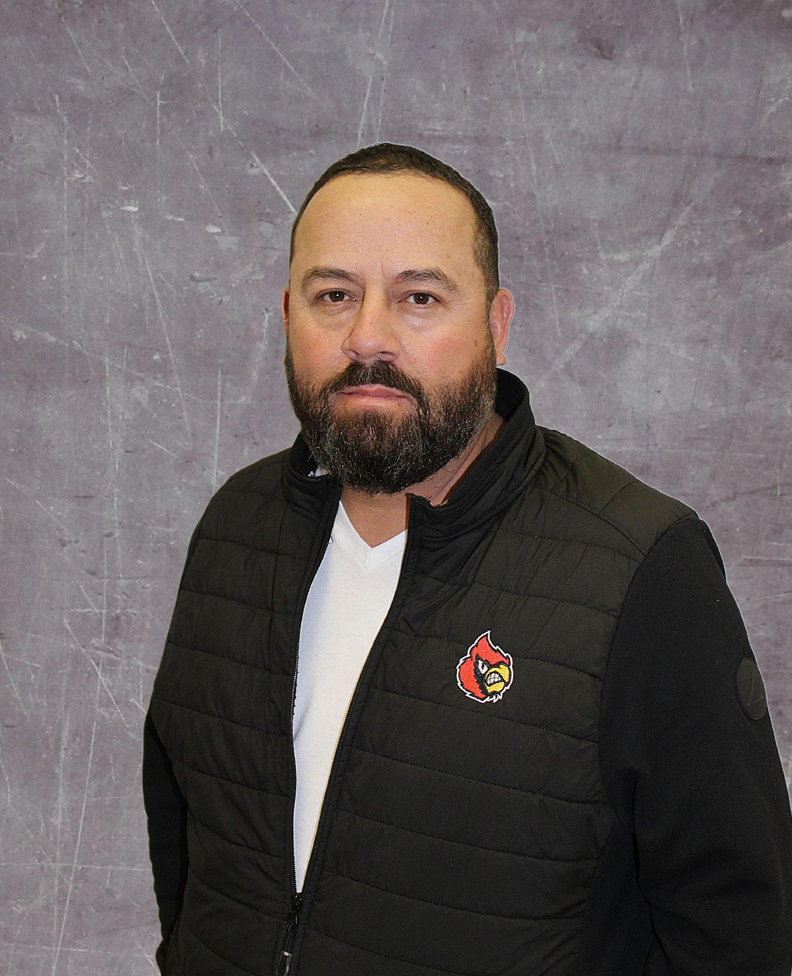 A man with a beard wearing a black vest with a logo, standing against a textured gray background.