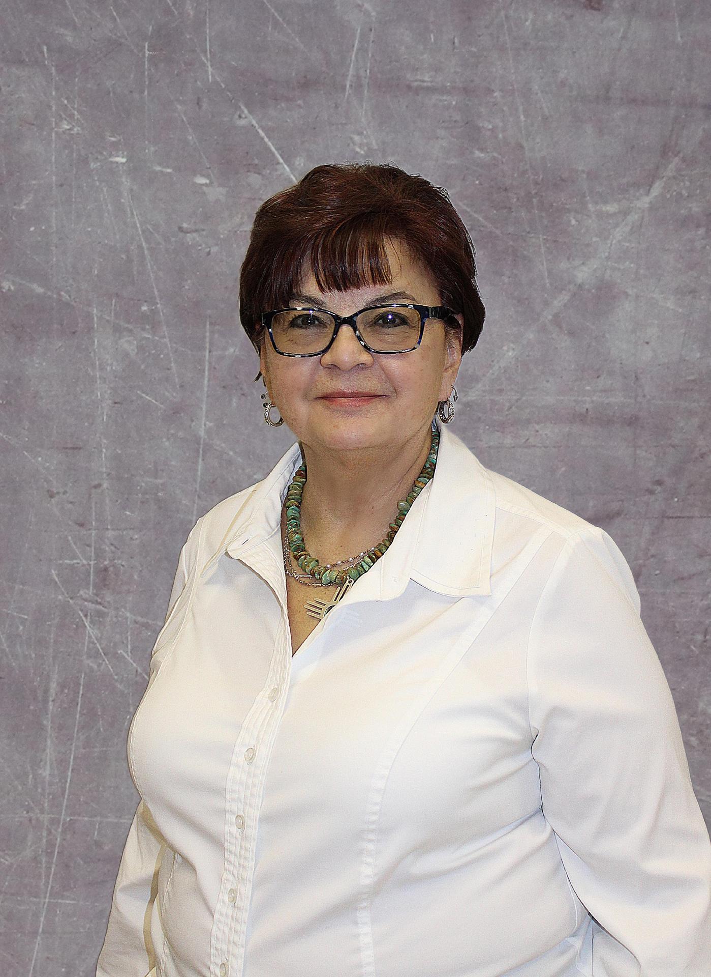 A woman with short brown hair and glasses, wearing a white blouse and a necklace, stands in front of a gray backdrop.