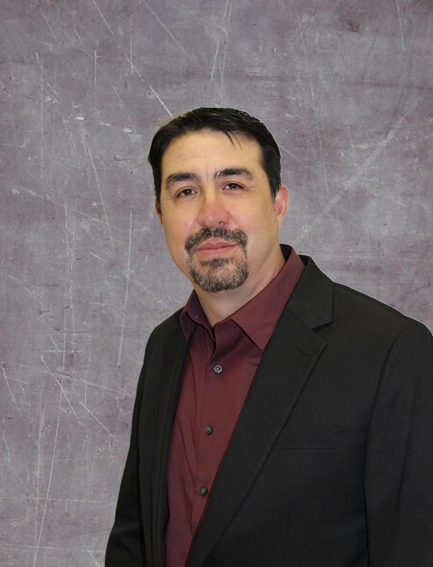 A man with dark hair and a goatee is wearing a black suit jacket over a maroon shirt, standing against a textured gray background.