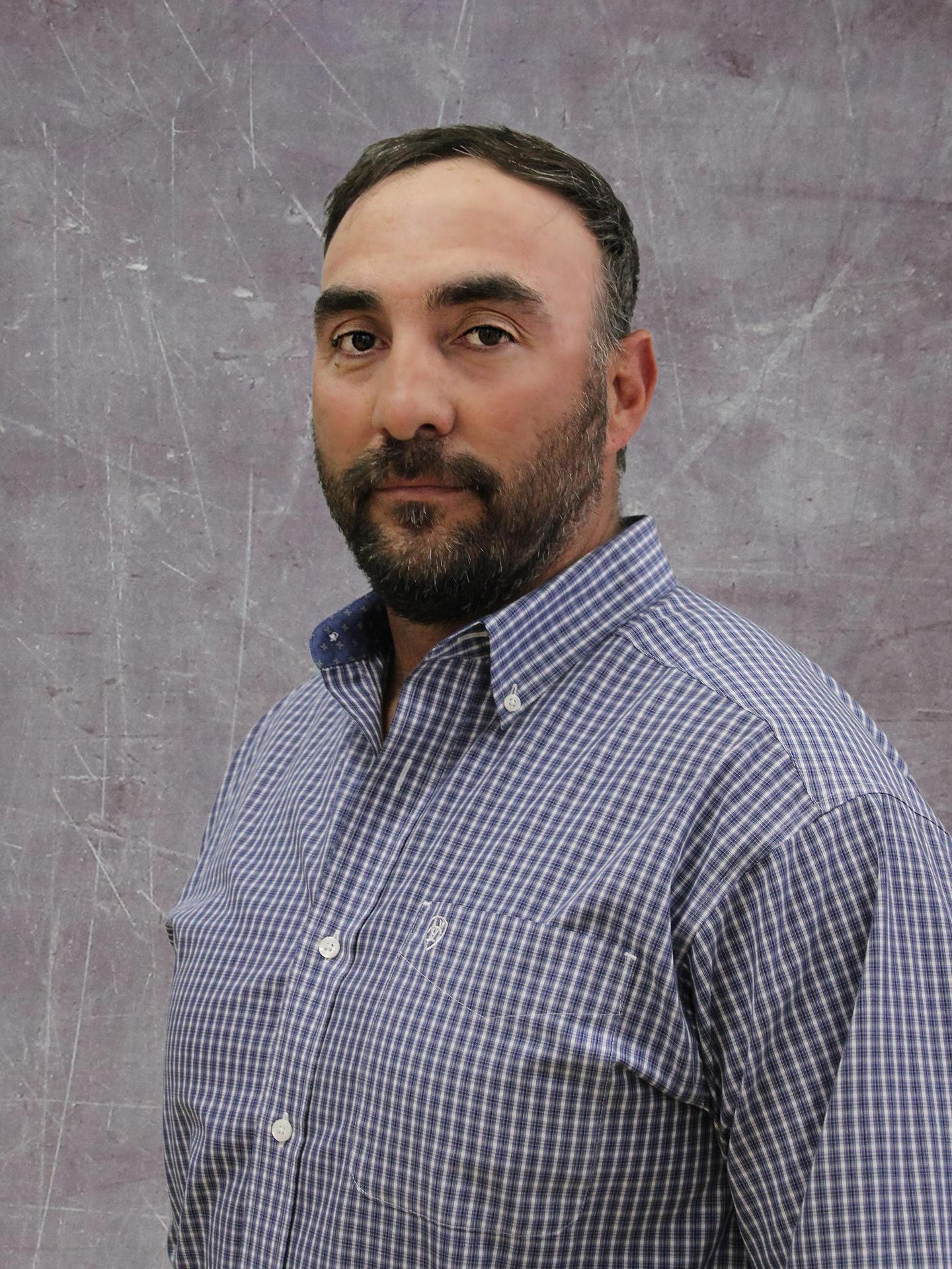 A man with a beard and short hair is wearing a checkered shirt, standing against a textured gray background.