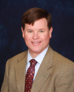 A man with short brown hair, wearing a tan blazer over a white shirt and a patterned tie, smiles against a dark blue background.