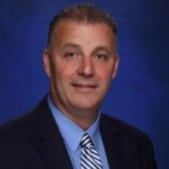 A professional headshot of a man in a suit with a light blue shirt and a striped tie, set against a blue background.
