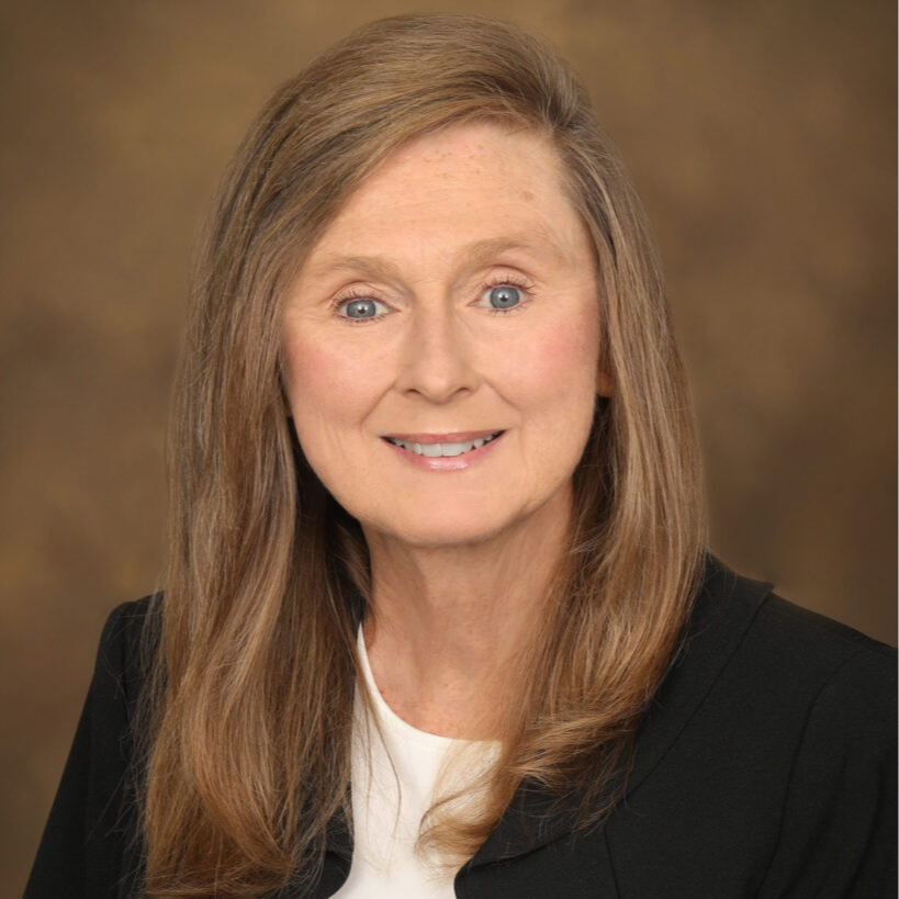 A professional headshot of a woman with long brown hair, wearing a black blazer over a white top, smiling against a blurred brown background.
