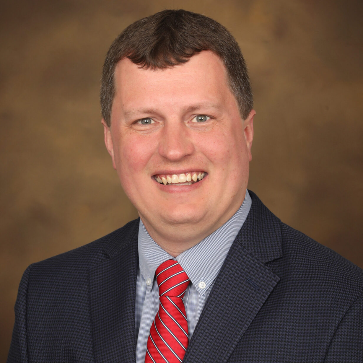 A smiling man in a dark suit and red tie poses for a professional headshot against a neutral background.