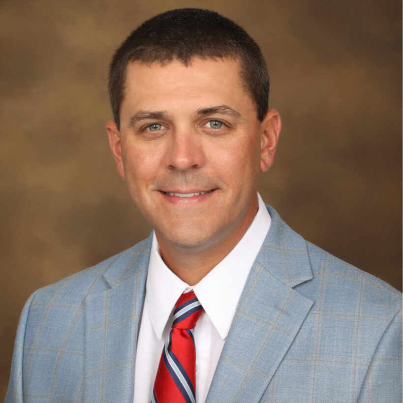 A man in a light blue suit with a white shirt and red and blue striped tie smiles at the camera against a blurred brown background.