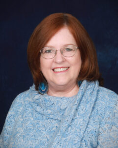 A woman with shoulder-length reddish-brown hair, wearing glasses and a blue sweater, smiles warmly at the camera against a dark blue background.