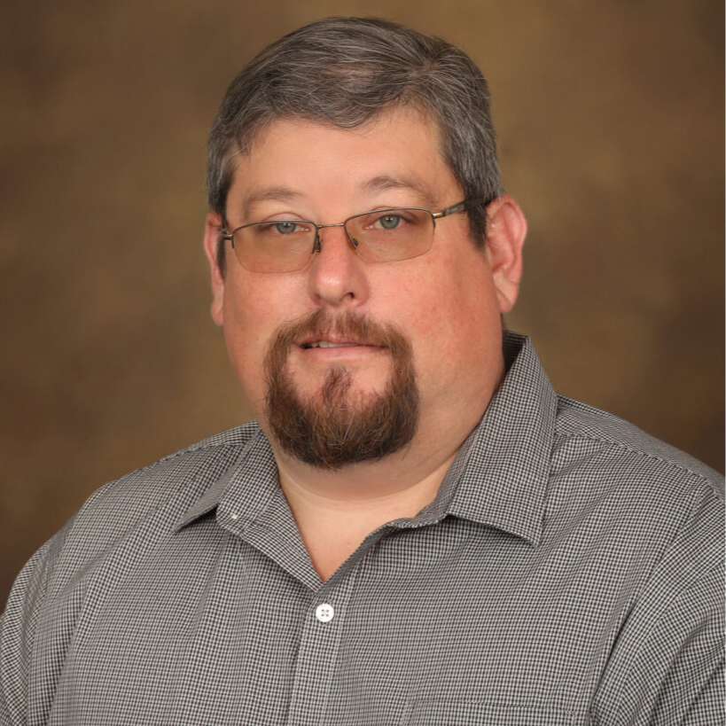 A man with glasses and a beard is wearing a gray checkered shirt, posing for a professional headshot against a blurred brown background.