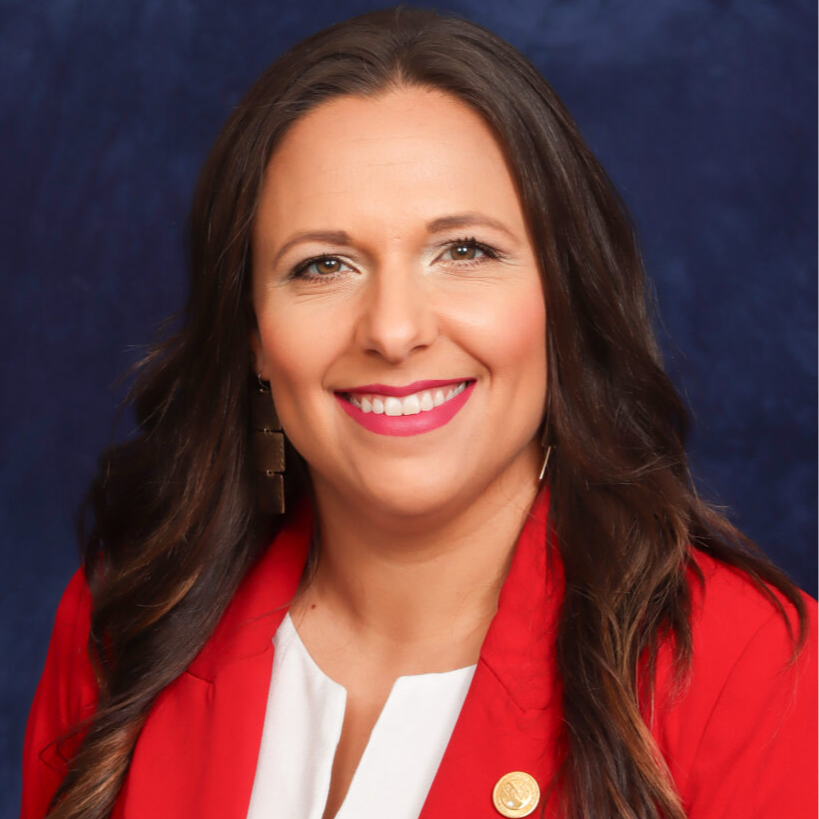 A woman with long, dark hair wearing a red blazer and white blouse smiles against a blue background.