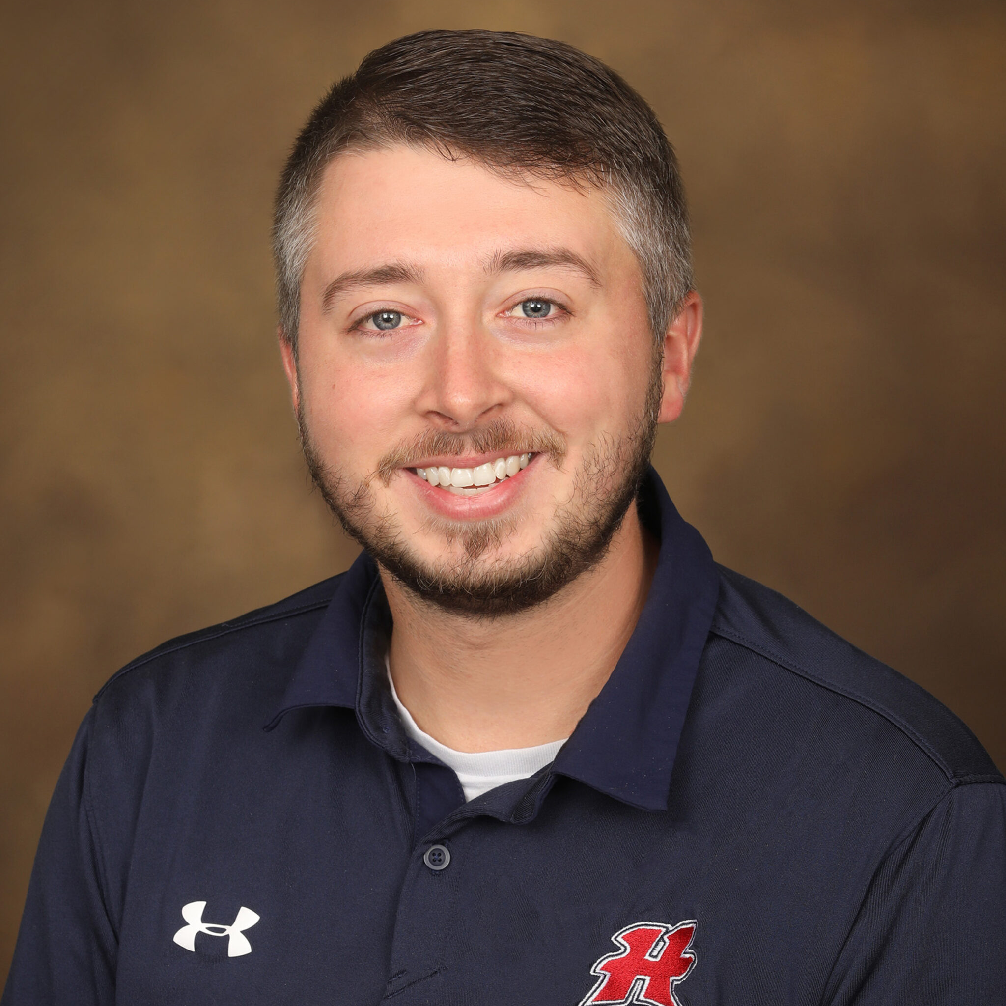 A smiling man with short hair and a beard is wearing a dark blue polo shirt with a logo on the left side, posing against a blurred brown background.