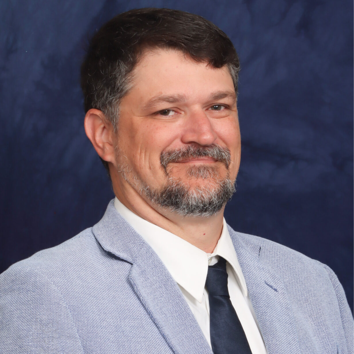 A man with a beard and mustache is smiling, wearing a light blue blazer over a white shirt and a dark tie, against a dark blue background.