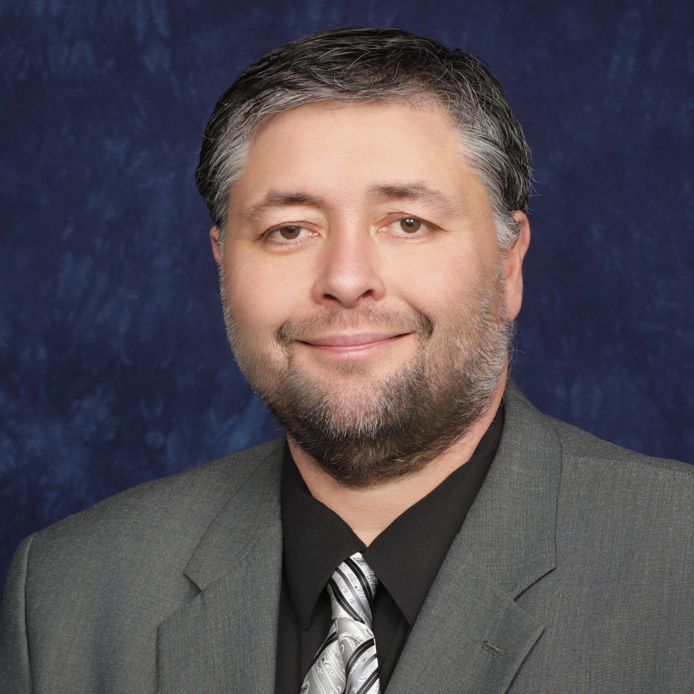 A man with a beard and short hair, wearing a gray suit and a red shirt with a tie, poses against a dark blue background.