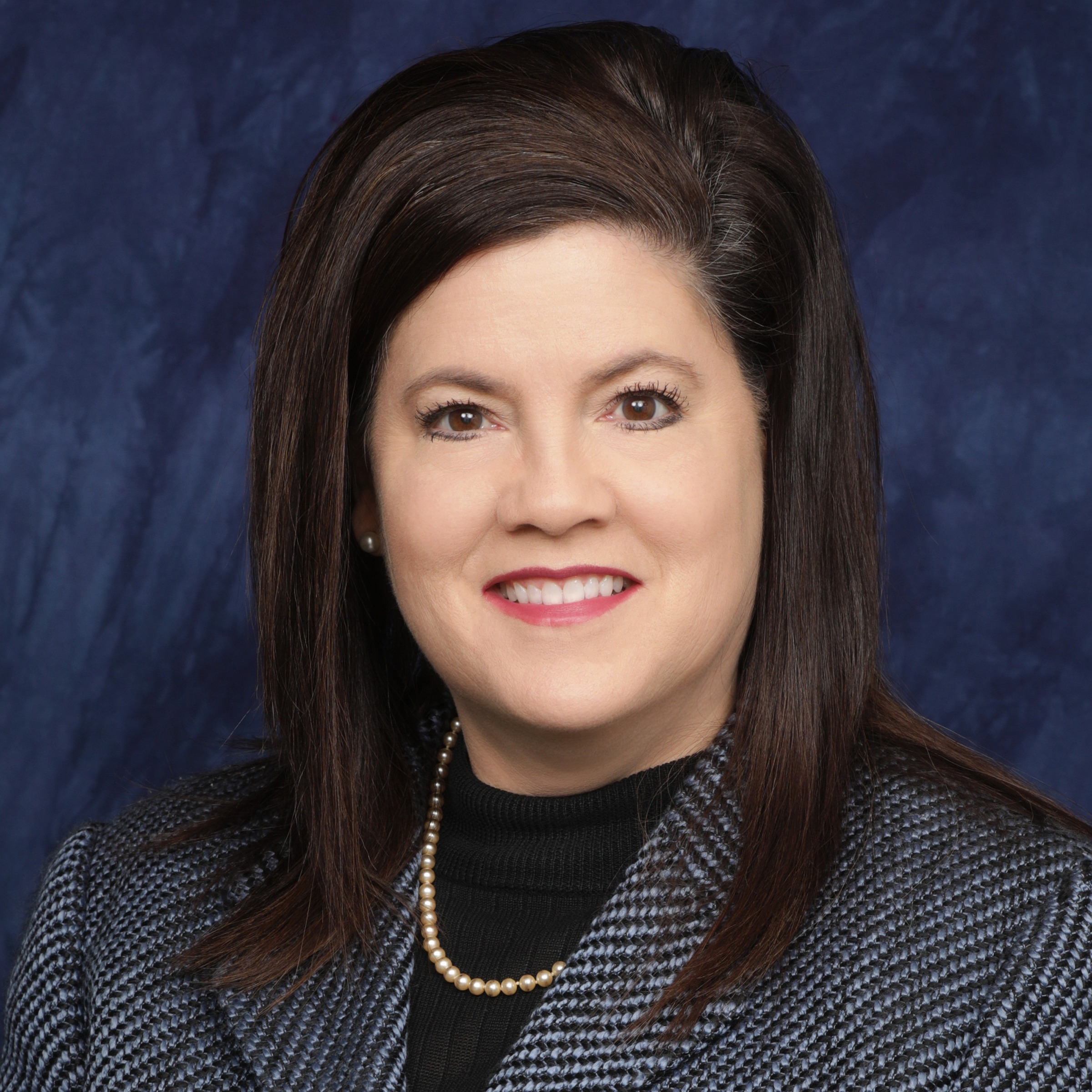 A woman with long brown hair wearing a light green blazer over a white blouse, smiling against a dark blue background.