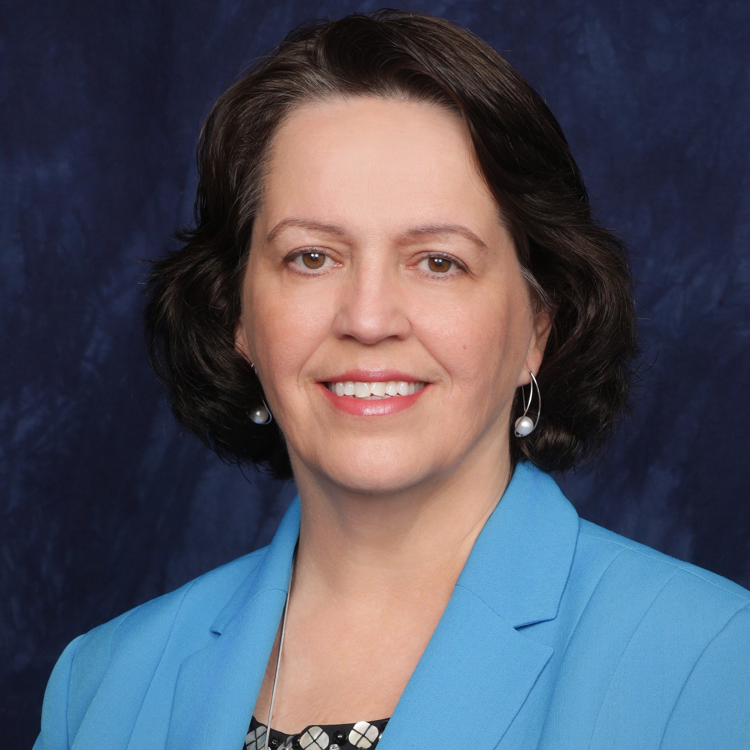 A woman with shoulder-length dark hair smiles warmly, wearing a maroon blazer over a patterned blouse, against a dark blue background.