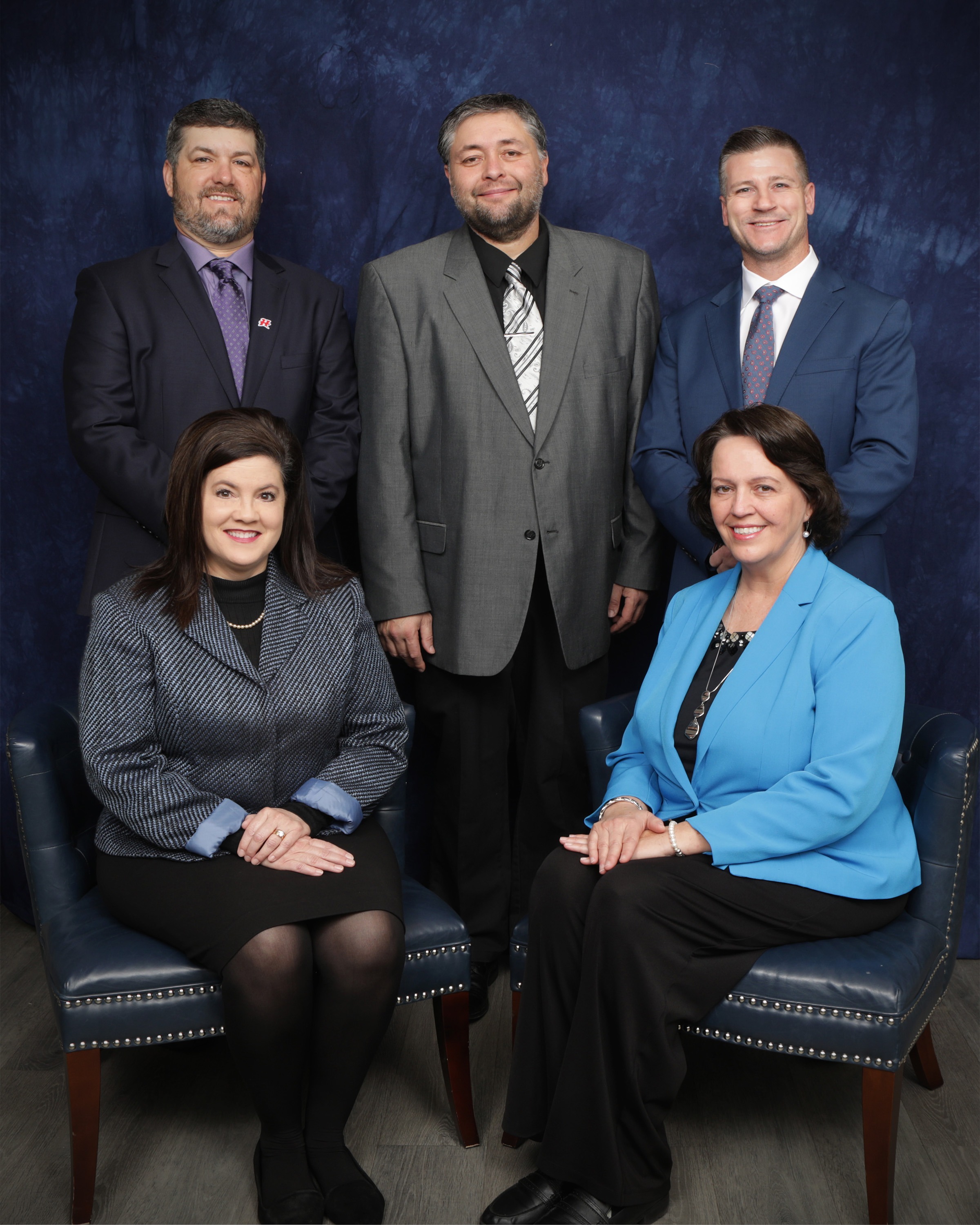 A group portrait of five individuals, with three men standing in the back and two women seated in front, all dressed in professional attire against a dark blue background.