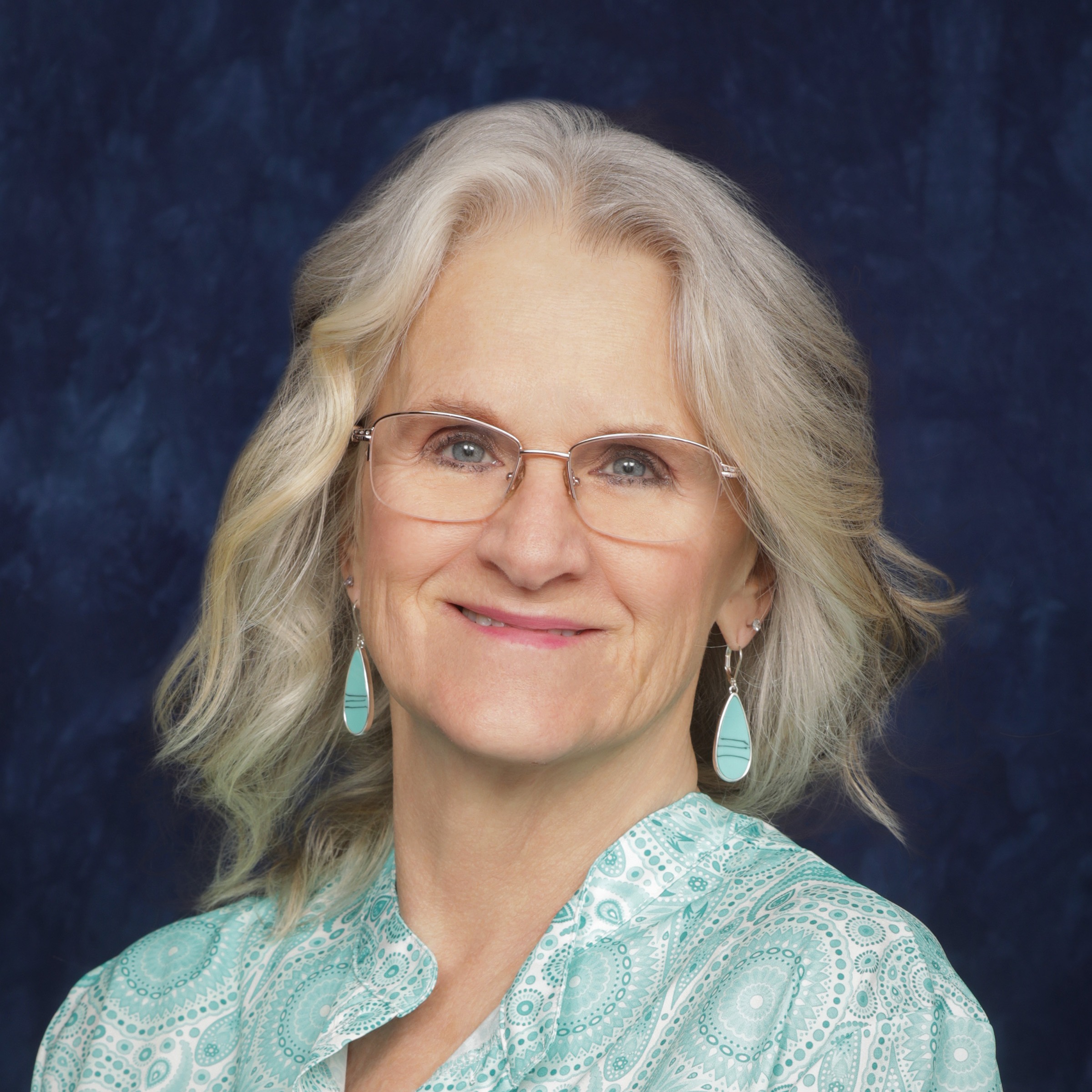 A woman with long blonde hair and glasses smiles at the camera, wearing a blue patterned top against a dark background.