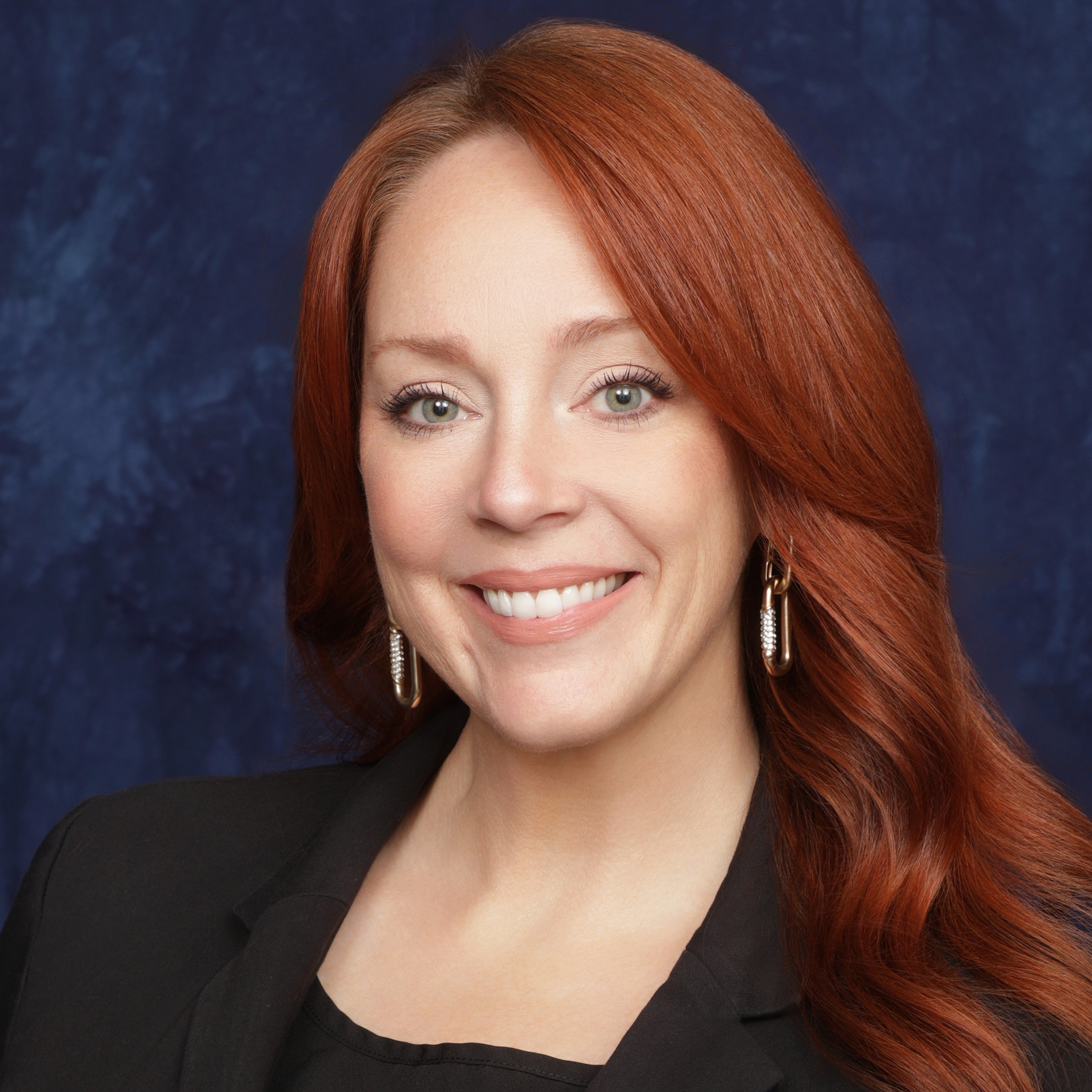 A smiling woman with long red hair, wearing a black top and a light-colored blazer, poses against a neutral brown background.