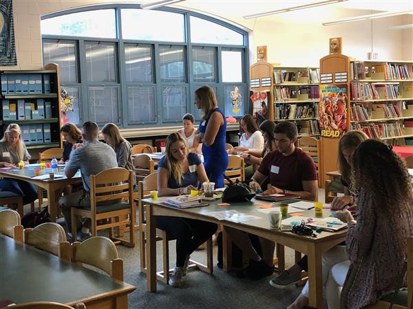 A group of people engaged in activities at tables in a library, with bookshelves in the background and large windows allowing natural light to enter.