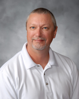 A man with short gray hair and a beard is wearing a white polo shirt, smiling at the camera against a soft gray background.